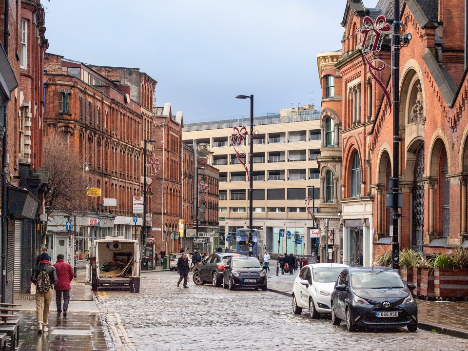 View of High Street in Manchester's Northern Quarter, featuring brick buildings, parked cars, and pedestrians.