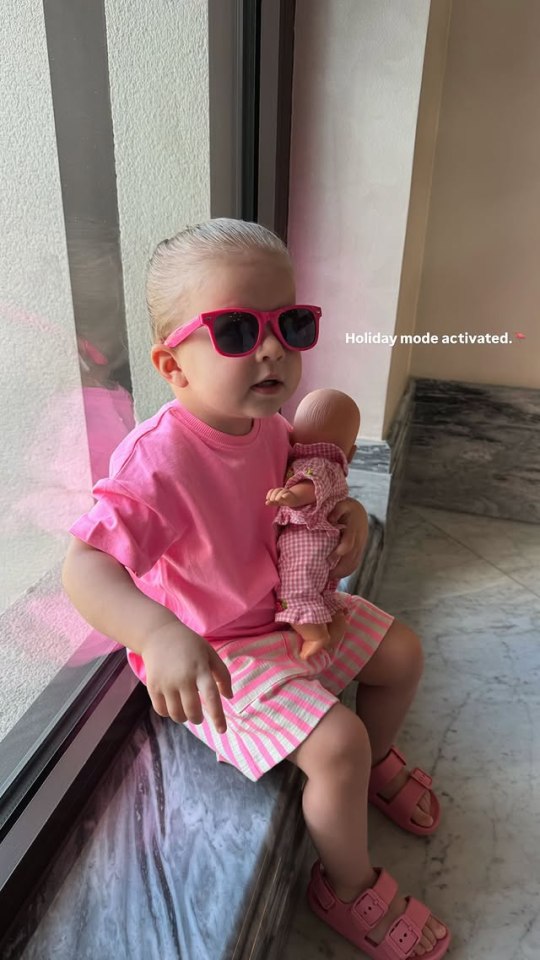 Toddler wearing pink sunglasses and holding a doll.