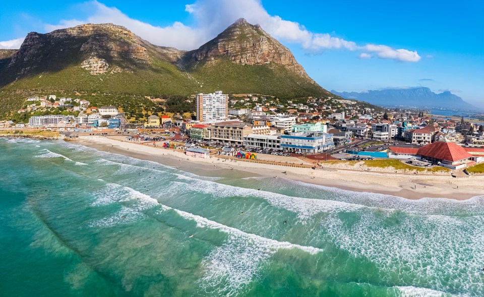 Aerial view of Muizenberg Beach in Cape Town, South Africa, showing surfers in the ocean and the town's buildings against a mountain backdrop.