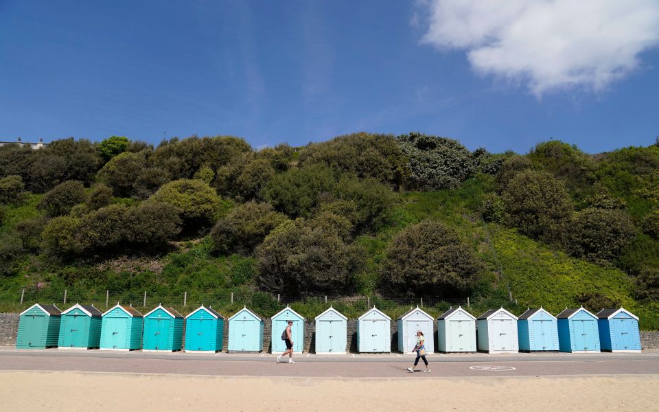 Two people walking past a row of beach huts on a sunny day.