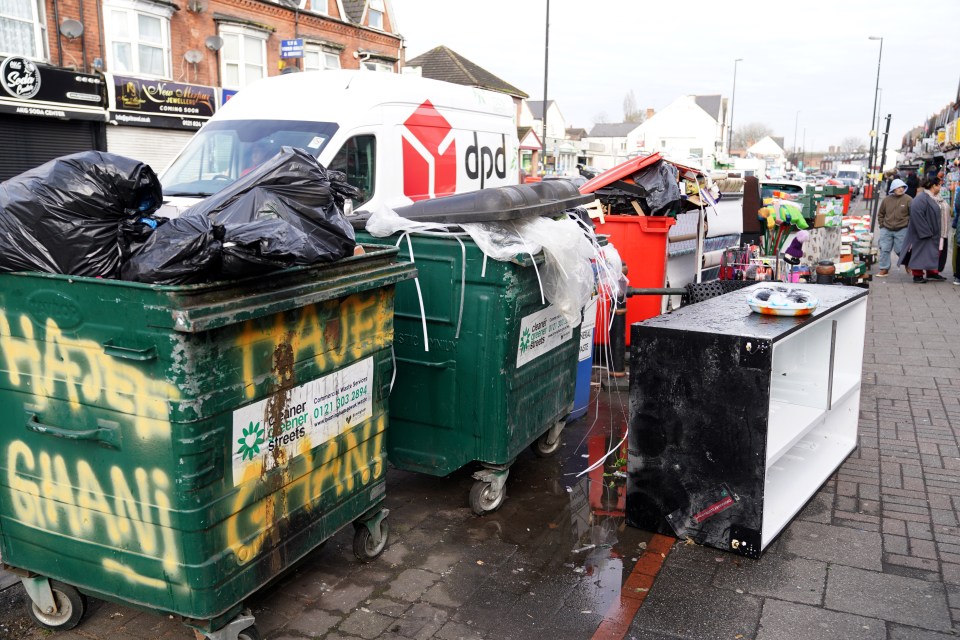 Overflowing wheelie bins and rubbish bags piled high on a city street.