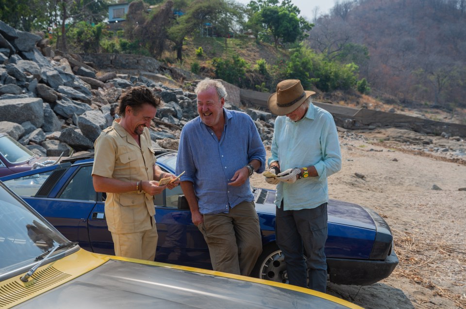 Three men standing by classic cars, looking at something in their hands.