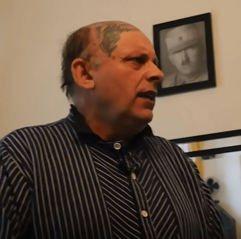 Man with a head tattoo speaking, with a framed portrait of Adolf Hitler visible in the background.
