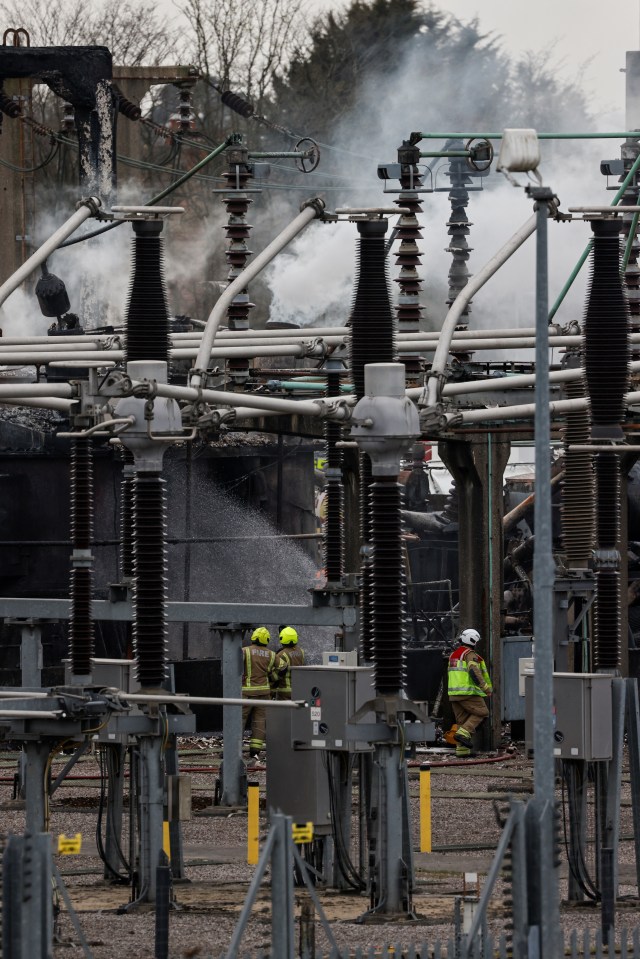 Smoke rises from a damaged electrical substation with firefighters present.
