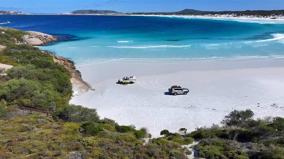 Aerial view of two vehicles parked on a white sand beach next to turquoise water.