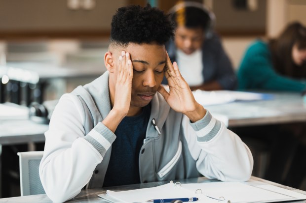 A stressed teenage boy holds his head in his hands while sitting at a desk.