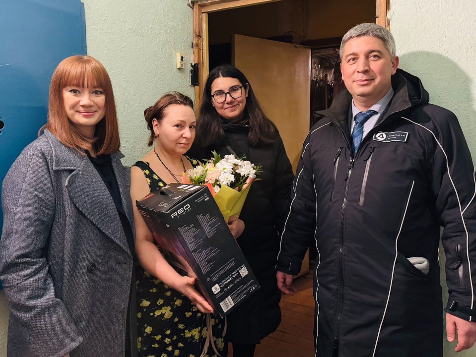 Photo of three women and a man; one woman is receiving a gift box and flowers.