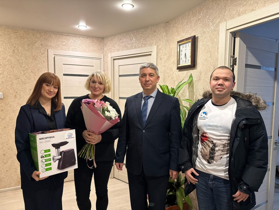 Photo of four people, two women and two men, one woman holding a meat grinder and a bouquet of flowers.
