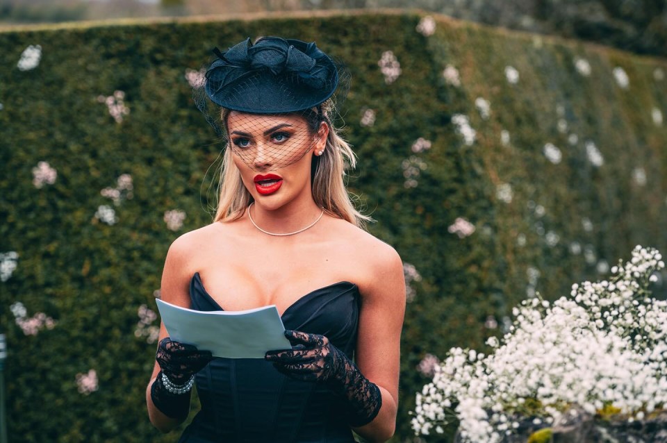 Woman in black dress and hat reading from a piece of paper.