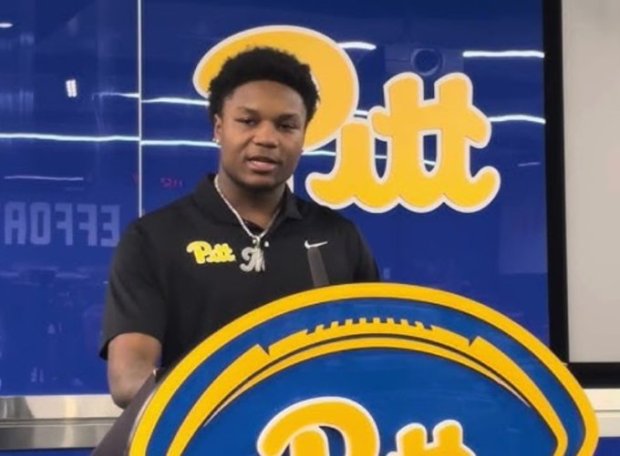 A young man in a Pitt Panthers shirt stands behind a Pitt football-shaped sign.