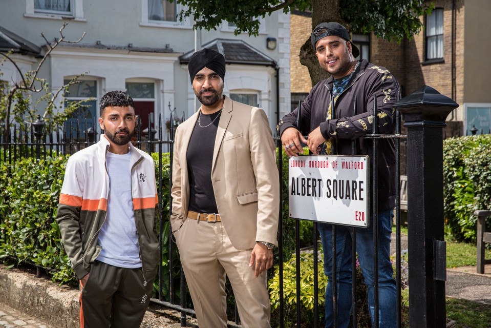 Three actors from EastEnders posing by an Albert Square sign.