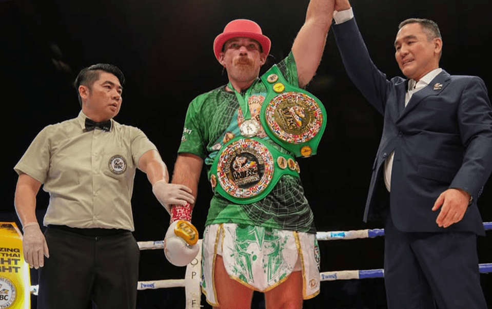 A boxer wearing a green shirt and a red hat holds up two championship belts.