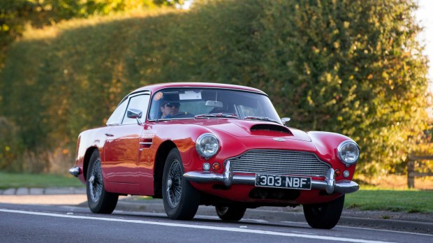 1960 red Aston Martin DB4 driving on a road.