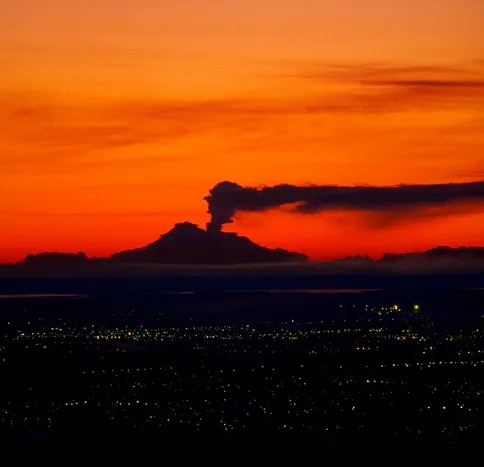 Mount Spurr volcano erupting at sunset, with city lights in the foreground.