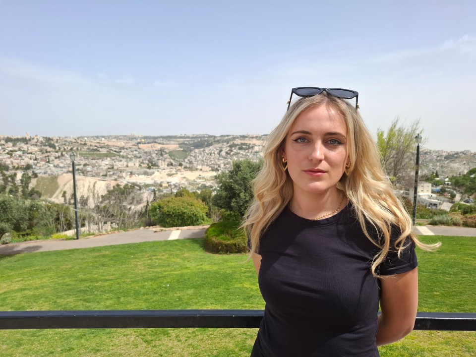 Woman with blonde hair and sunglasses in front of a city landscape.