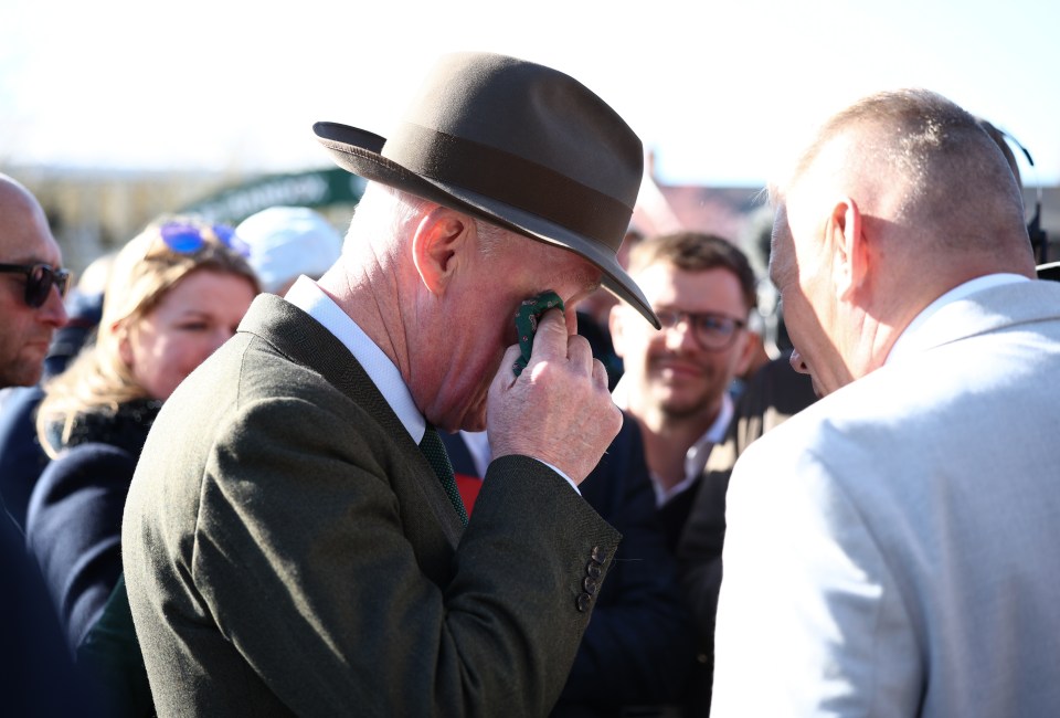 A man in a hat wipes his eyes while talking to another man.