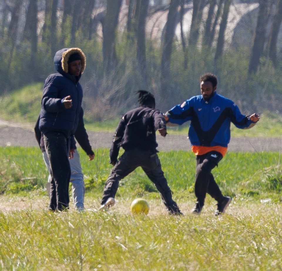 Migrants playing soccer in a field near an industrial plant.