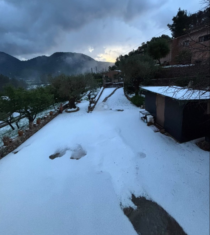 Hail covering a yard and pathway in front of a house and mountains.