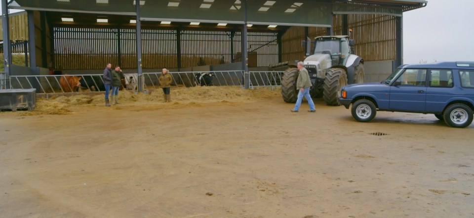 Three people standing near cows in a barn with a tractor and Land Rover parked nearby.
