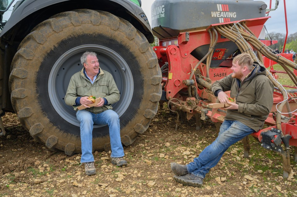 Two men eating sandwiches while sitting near farm equipment.
