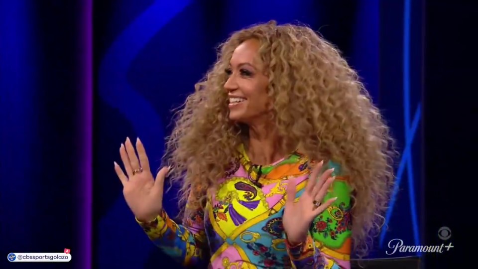 Woman with long curly hair, wearing a colorful patterned dress, gestures with both hands.
