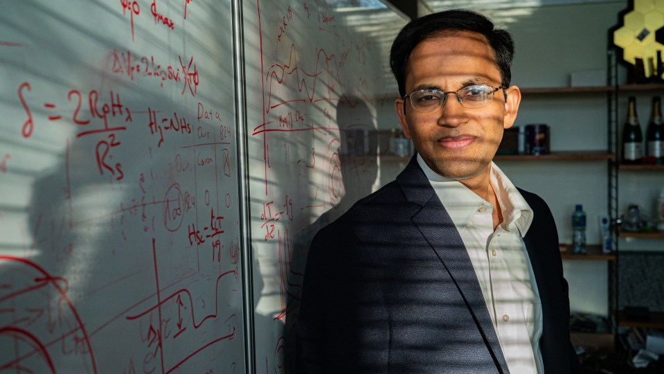 Portrait of Nikku Madhusudhan standing near a whiteboard with equations.