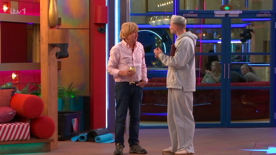 Screenshot of two men talking in a brightly lit room, with other people visible through a glass door in the background.