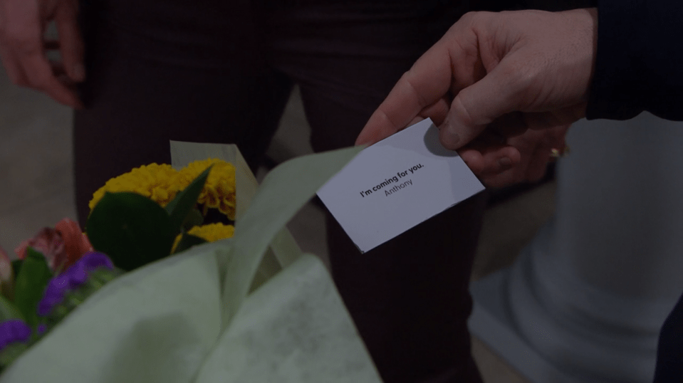 A hand holding a note that says "I'm coming for you. Anthony" above a bouquet of flowers.