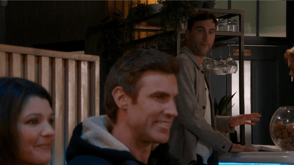 Screenshot of three people at a bar; two are seated, one is standing.