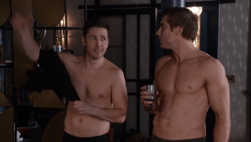 Screenshot of two shirtless men talking; one is taking off a shirt.