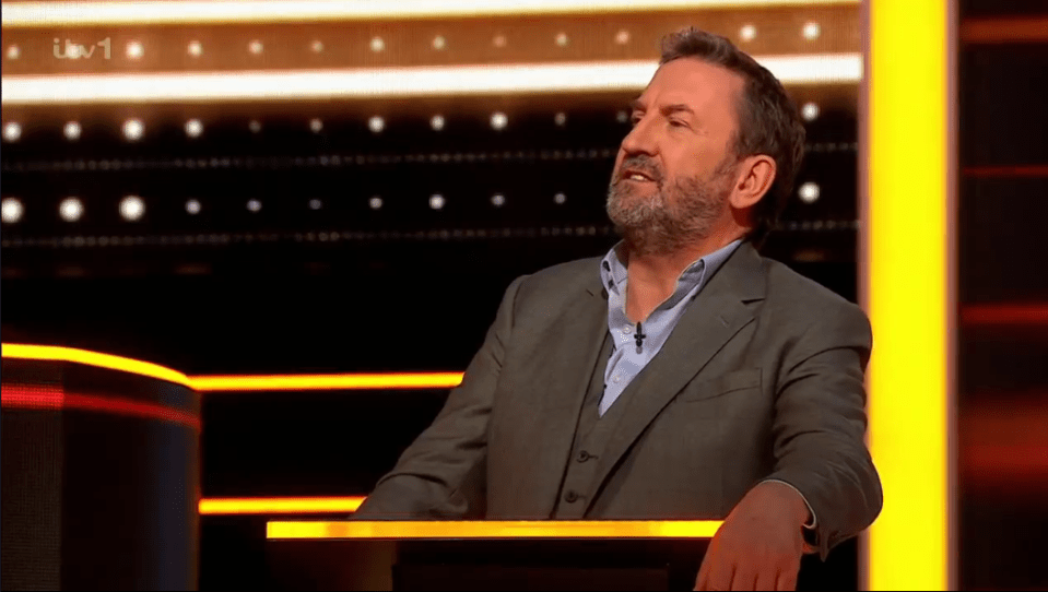 Screenshot of a man in a suit sitting at a game show podium.