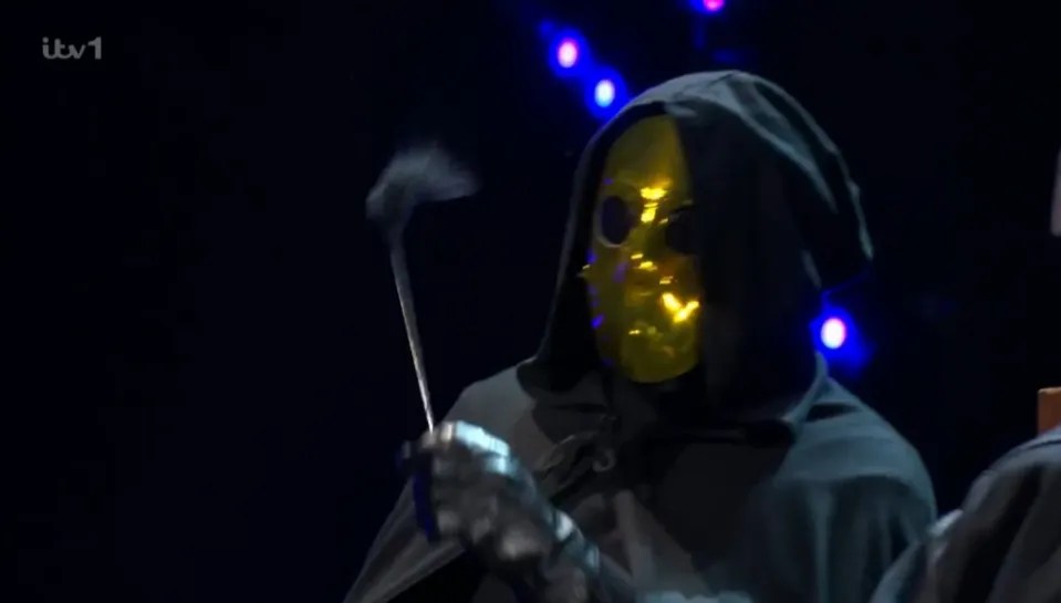 Screenshot of a person in a dark hooded robe and gold mask holding a smoking object.