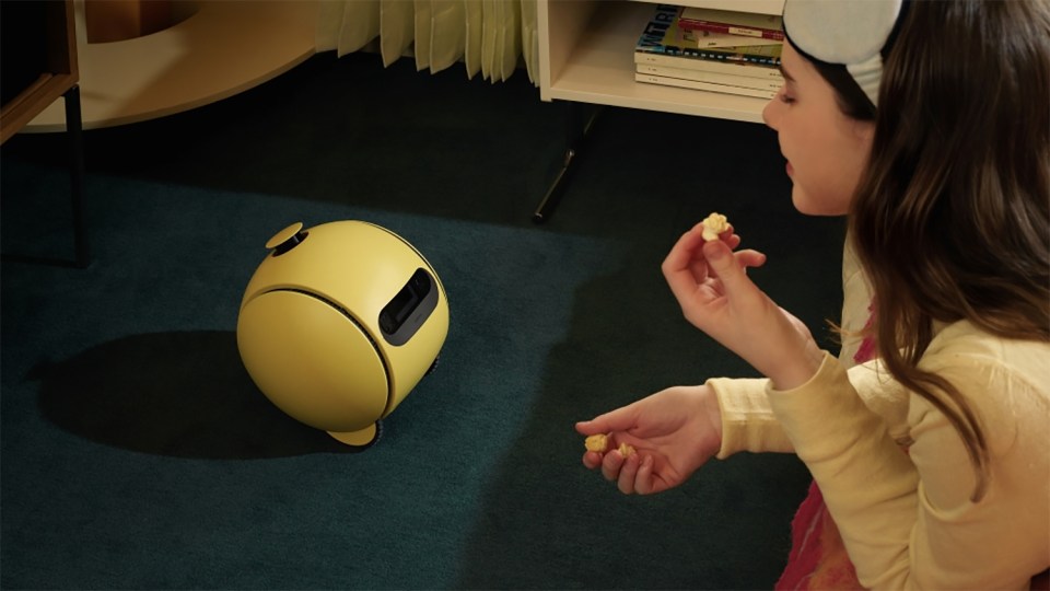 A yellow robot on the floor in front of a girl eating popcorn.