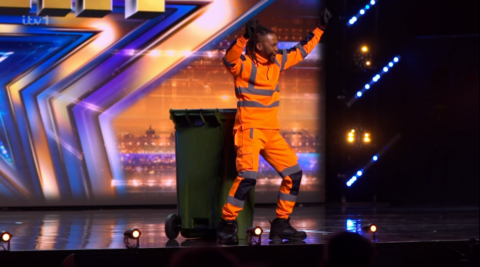 A man in an orange jumpsuit and safety vest dances next to a large green trash can on a stage.