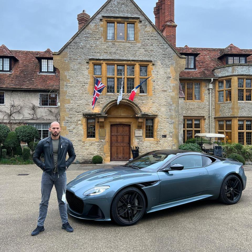 Man standing in front of a gray Aston Martin car parked in front of a large house.