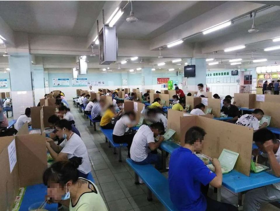 Workers eating lunch at individual desks in a Chinese factory cafeteria.