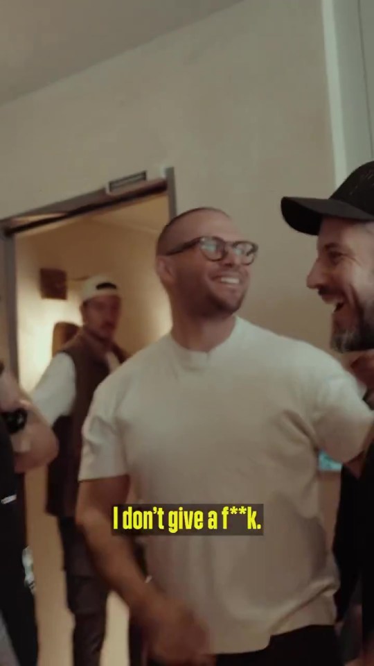 A man in glasses says "I don't give a f***."  Two other men are in the background.