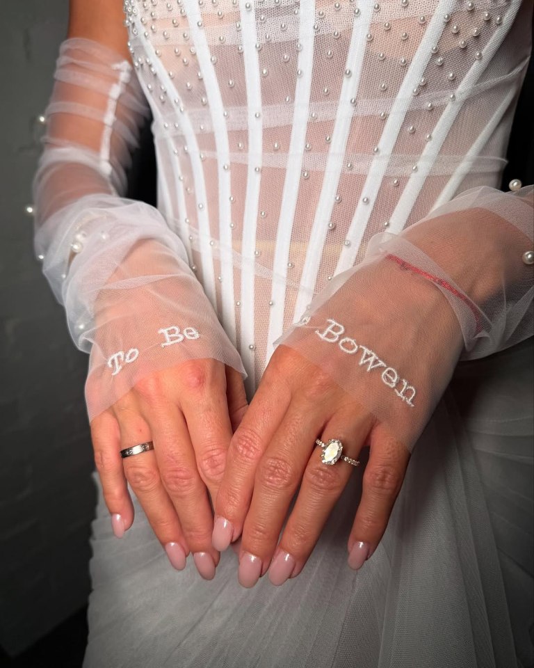 Close-up of hands wearing sheer gloves embroidered with "To Be" and "Bowen," showcasing an engagement ring.