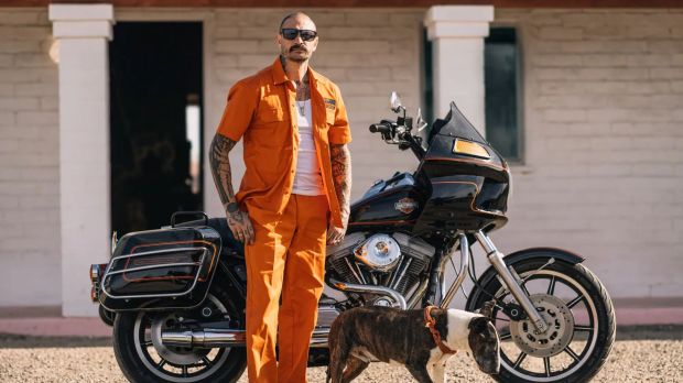 Man in orange Dickies jumpsuit standing with a dog next to a motorcycle.