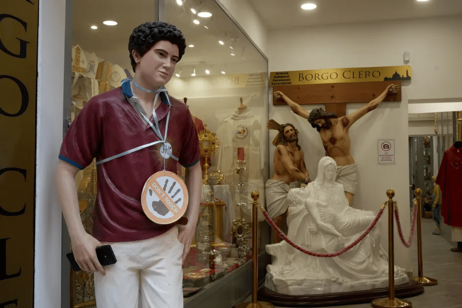Statue of a young man with a "Don't Touch" sign, displayed in a shop window with religious statues.