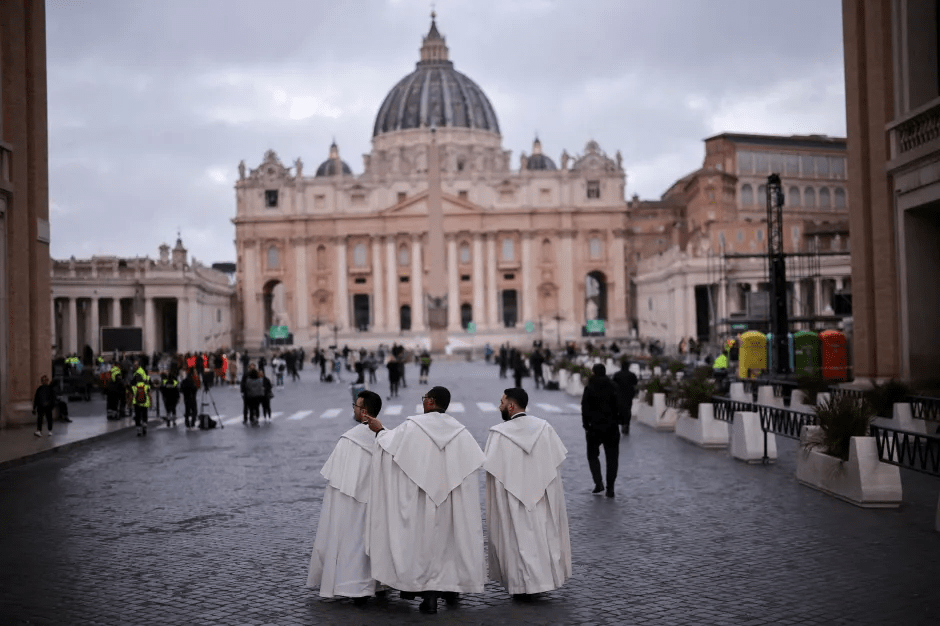 Three men in white robes walk away from the camera toward St. Peter's Basilica.
