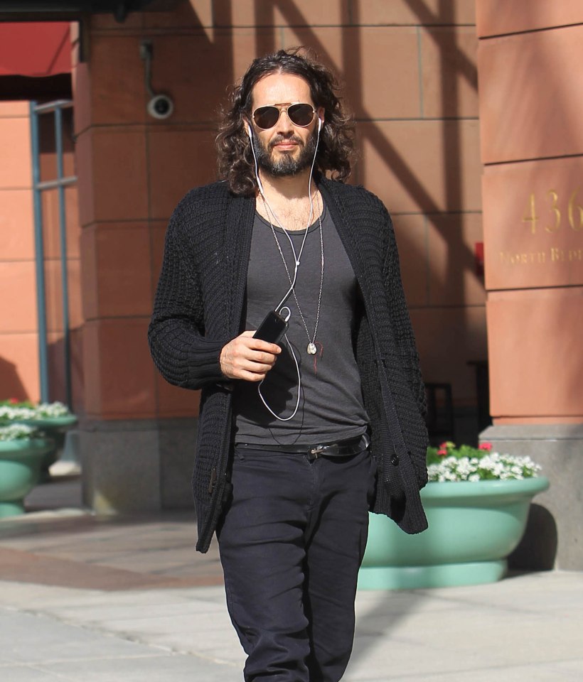 Russell Brand walking down the street, wearing sunglasses and earphones.