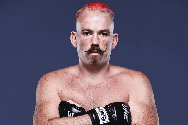 Portrait of a shirtless man with red hair, a handlebar mustache, and boxing gloves.