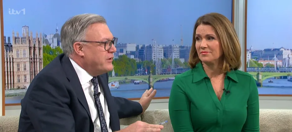 Two news presenters sitting on a sofa, discussing a topic.  A London cityscape is visible in the background.