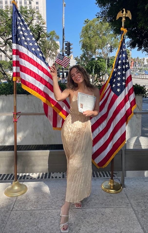 Woman in beige dress holding citizenship certificate between two American flags.