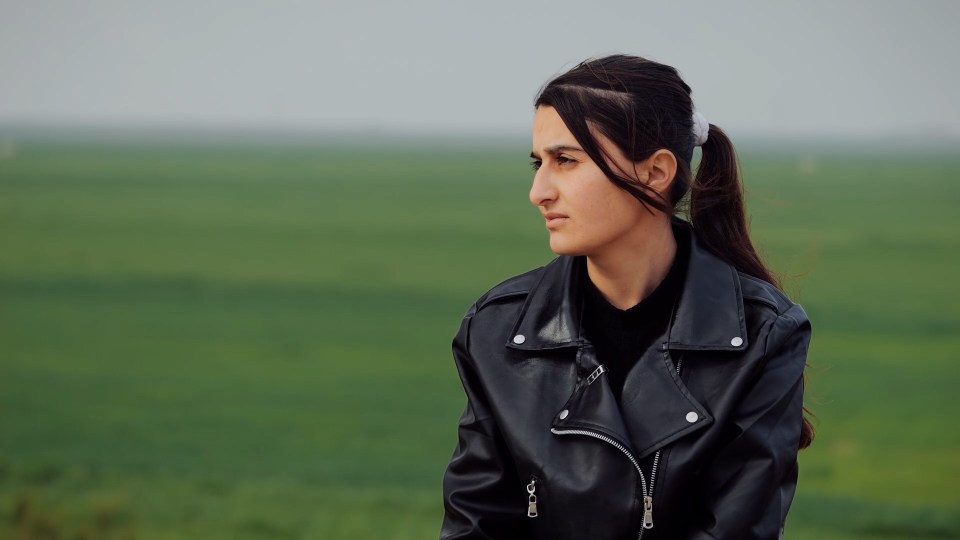 A young woman in a black leather jacket looks off to the side, against a blurred background of green fields.