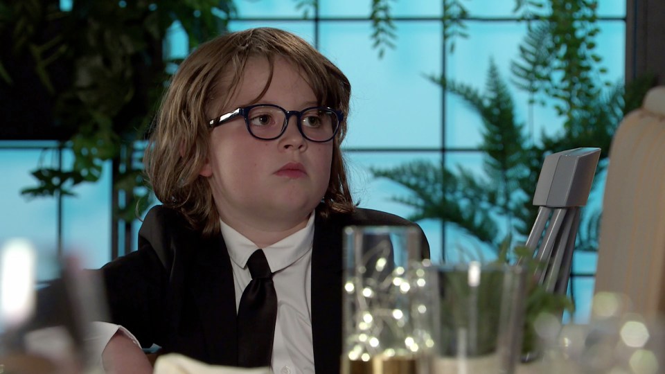 A boy in a suit and glasses sits at a table, looking serious.