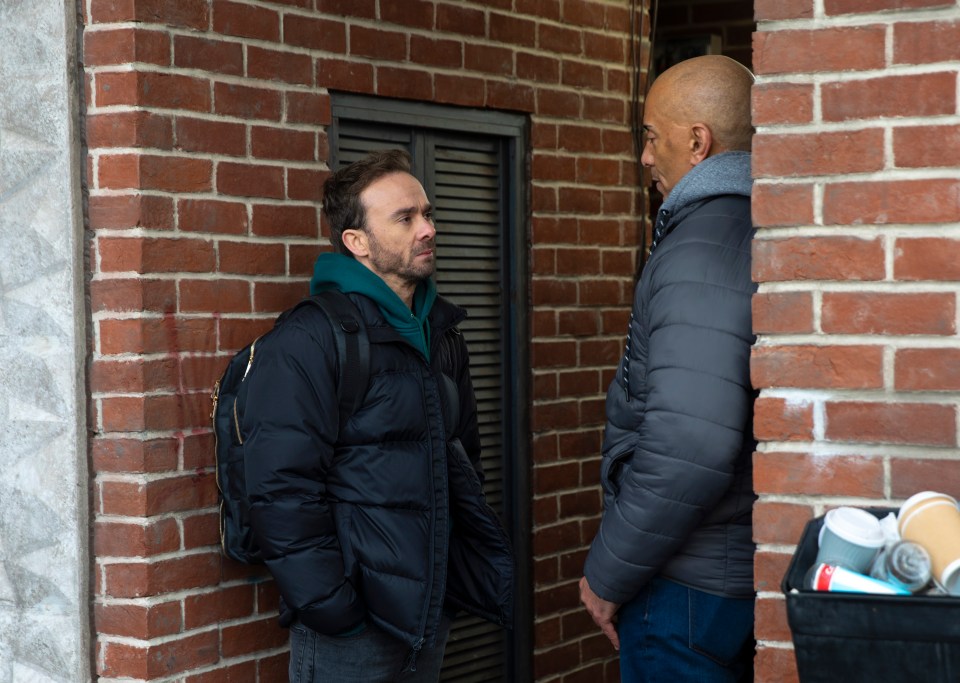 Two men talking against a brick wall.