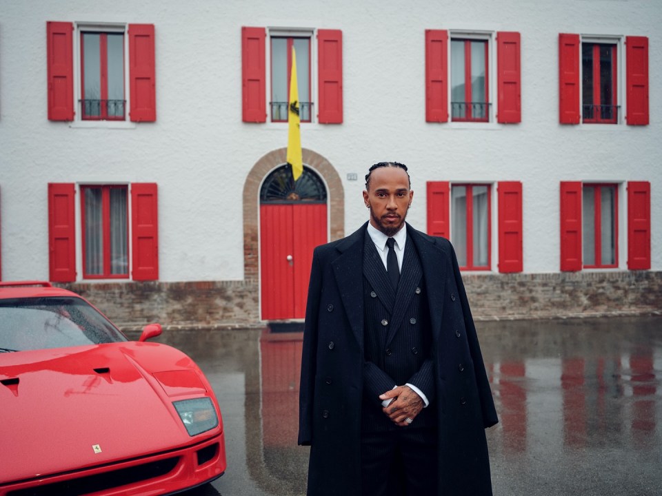Lewis Hamilton in a black suit and overcoat standing in front of a red Ferrari.