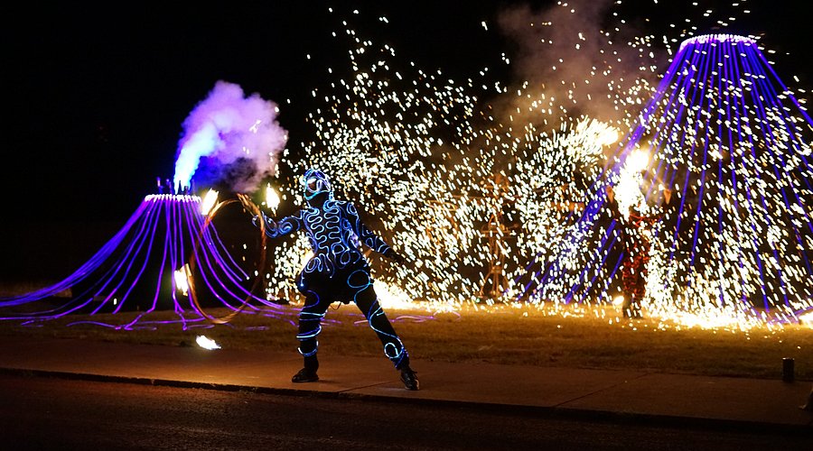 A performer in a light-up suit performs fire dancing in front of a volcano-shaped light display and fireworks.